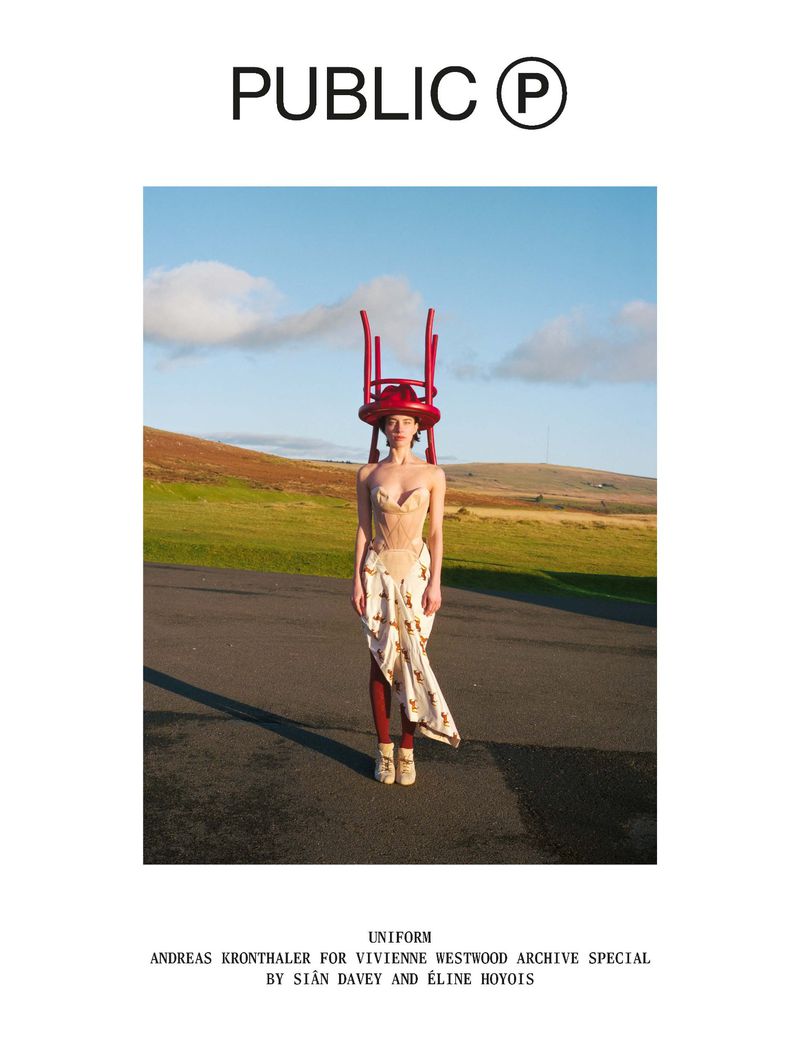 Model standing outdoors on pavement with a red chair balanced upside down on her head, set against a rolling moorland landscape.