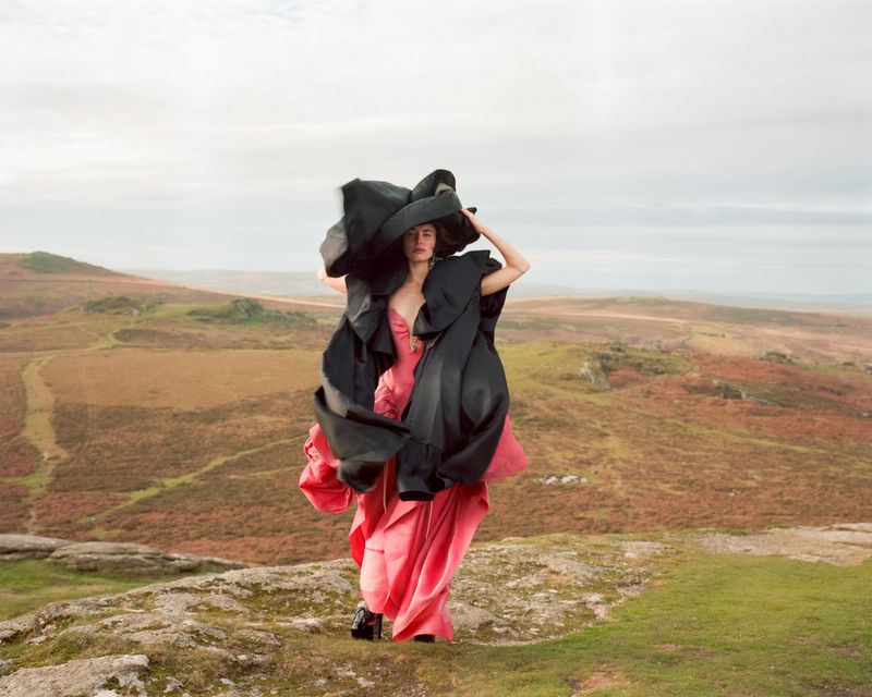Model standing on a moor wearing a voluminous black sculptural garment over a pink dress under an overcast sky.