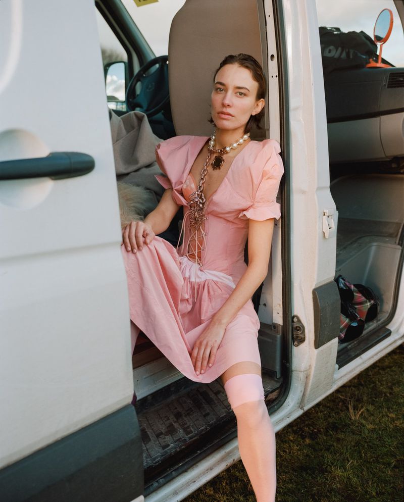 Woman in pink dress seated in the open doorway of a white van, wearing a pearl necklace and sheer pink stockings.