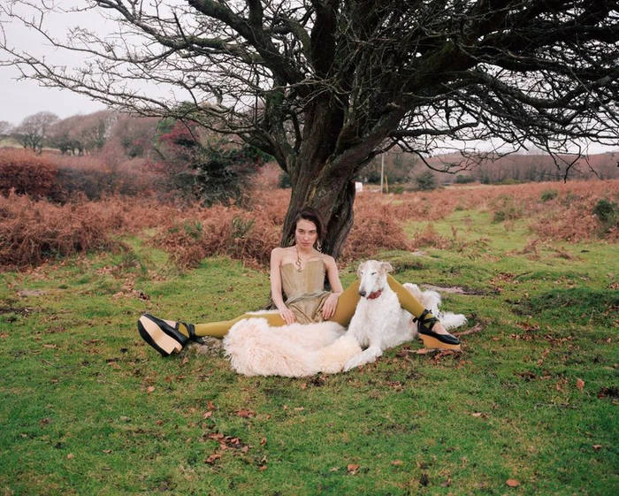 Model seated beneath a bare tree on a moor next to a large white borzoi dog, wearing a metallic corset and wedge platforms.
