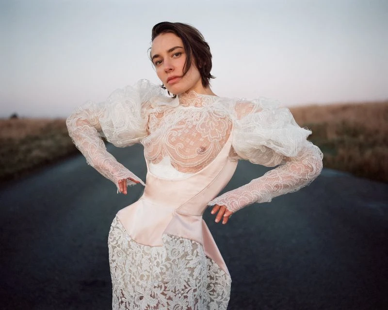 Model wearing a white lace dress with sheer puff sleeves standing on an empty outdoor road under an overcast sky.