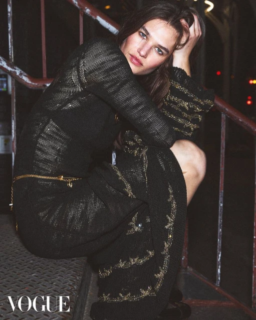 Fashion shot of an adult female model in a knit dress with embellishments, posing on a metal staircase in an urban environment at night.