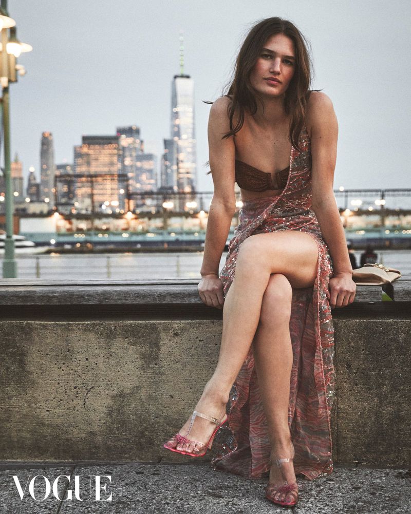 Fashion shot of a woman in an evening gown sitting on a concrete ledge with a cityscape backdrop, clear heels and Vogue logo.