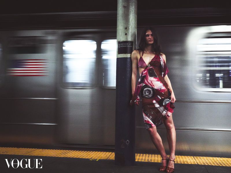 Fashion editorial shot of a woman in patterned dress leaning on a pillar at a subway station as a train car speeds by.