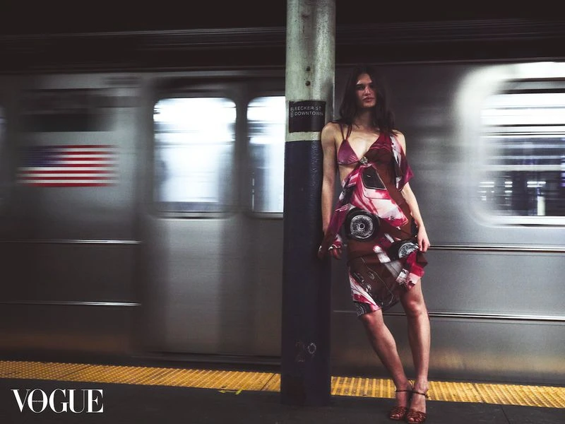 Fashion editorial shot of a woman in patterned dress leaning on a pillar at a subway station as a train car speeds by.