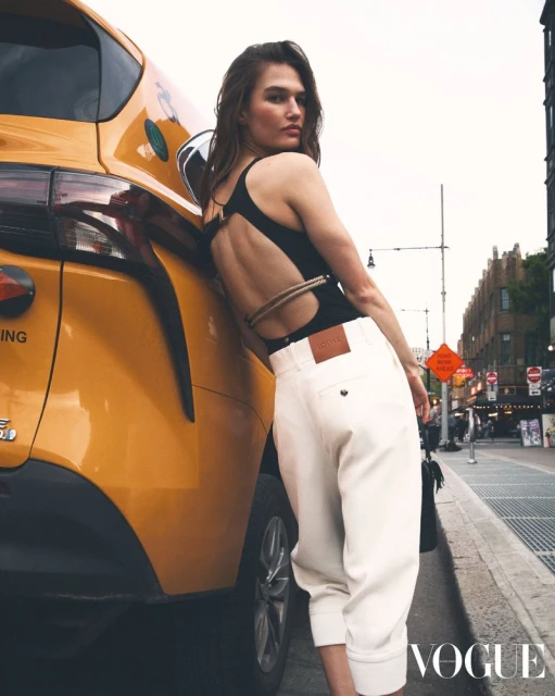 Adult female fashion model in black bodysuit and white pants leans against a yellow taxi in an urban setting, Vogue logo in corner.