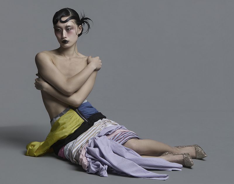 Model seated on a grey studio floor wearing layered multicolored garments and glitter heels for Vingt Sept Magazine.