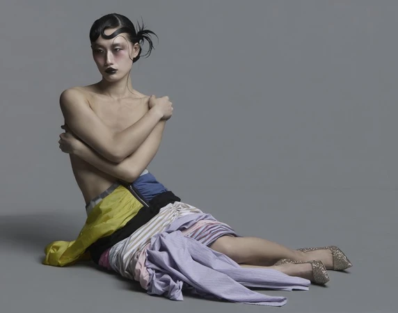 Model seated on a grey studio floor wearing layered multicolored garments and glitter heels for Vingt Sept Magazine.