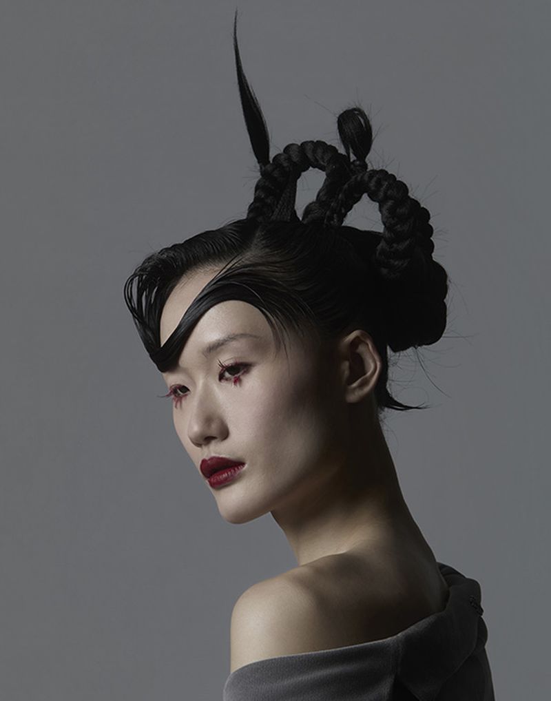 Close-up portrait of an East Asian woman with avant-garde braided black hair and dramatic red lipstick against a plain grey background