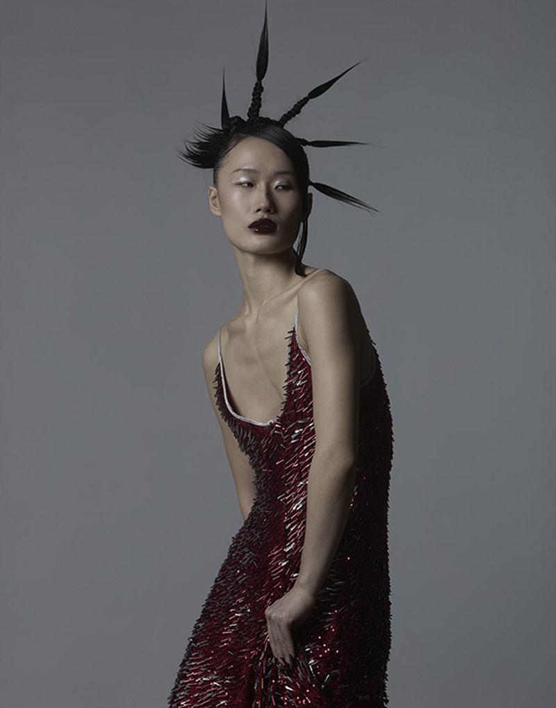 Model with spiky avant-garde hair wearing a dark red embellished dress against a plain grey backdrop.