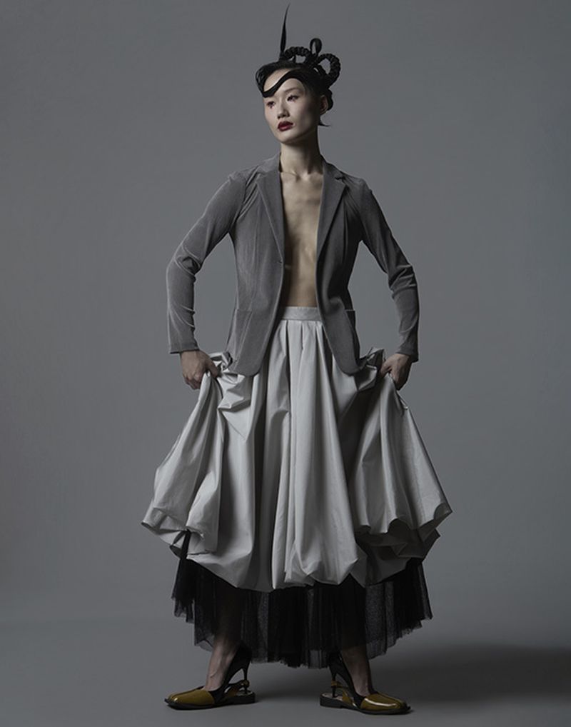 Model wearing a grey blazer, voluminous pleated skirt, and avant-garde sculpted hair against a grey background.