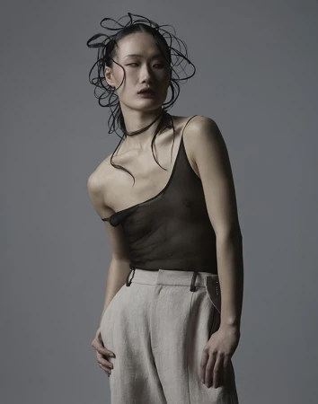 Asian woman wearing a sheer black top and beige trousers with sculptural dark hair against a grey background