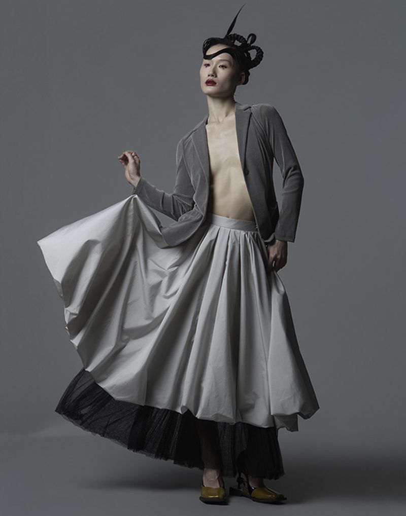 Fashion portrait of an adult woman wearing a grey blazer, full skirt, and artistic headpiece, posing in a photo studio.