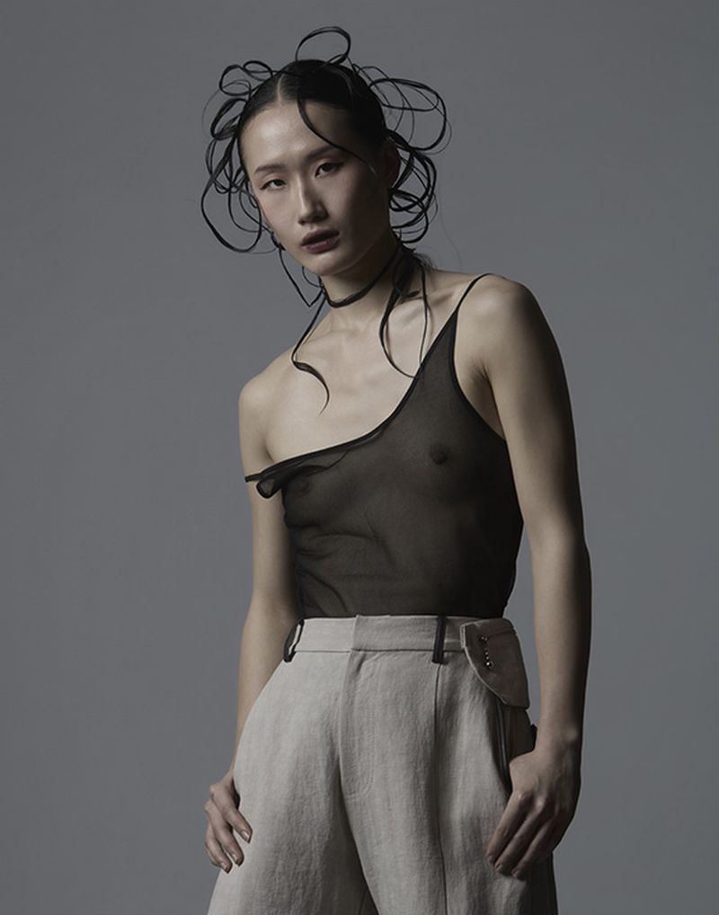 Model in sheer black top and beige trousers with intricate hair styling against a plain grey studio background.