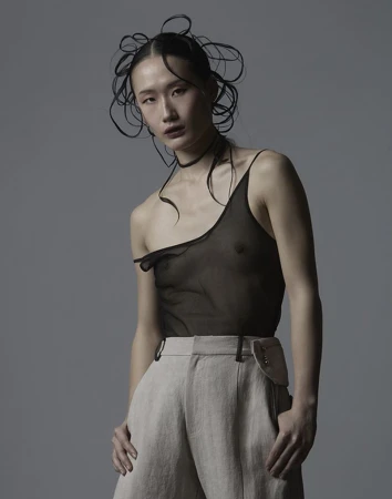 Model in sheer black top and beige trousers with intricate hair styling against a plain grey studio background.
