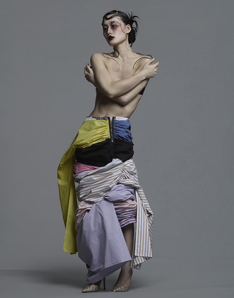 A model with dark wet-look hair and bold makeup wearing a layered colorful fabric skirt against a grey background.
