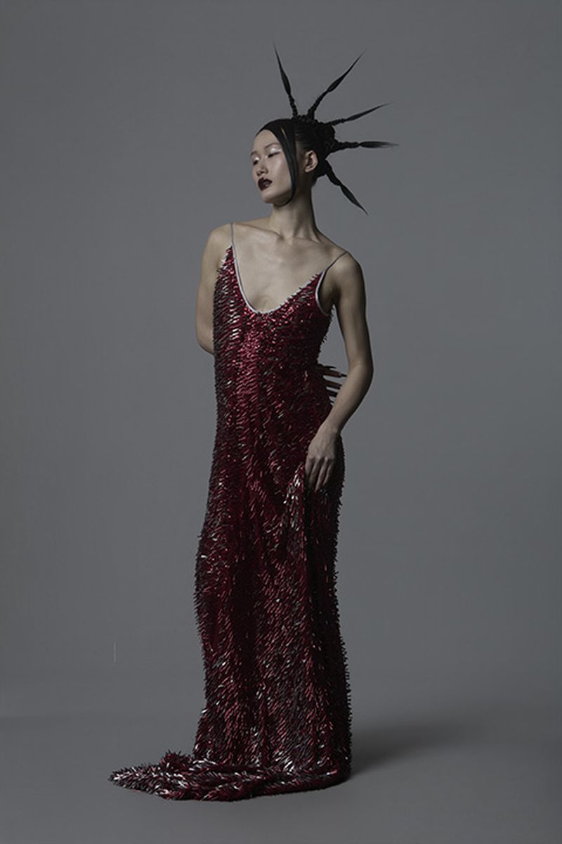 Model in a textured red sequin dress with spiky black hair against a neutral grey studio backdrop for Vingt Sept Magazine