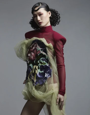 Asian woman wearing a red mock-neck top and sheer green draped dress against a grey backdrop for Vingt Sept Magazine