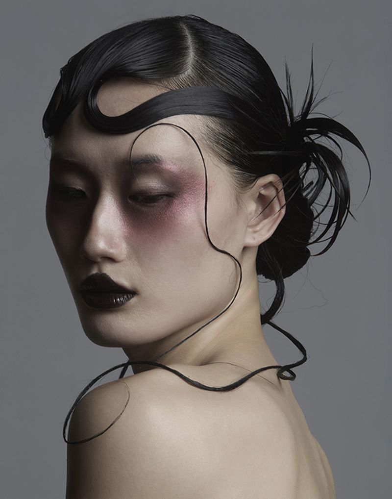Close-up portrait of a woman with slicked black hair, sculpted hair waves, deep red eyeshadow, and dark glossy lips against grey.
