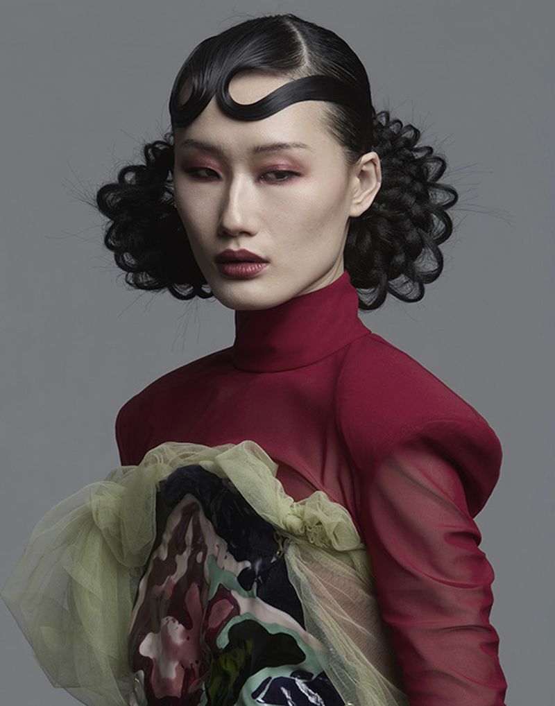 Close-up portrait of an East Asian woman with avant-garde sculpted dark hair and high-neck red sheer top against a grey background