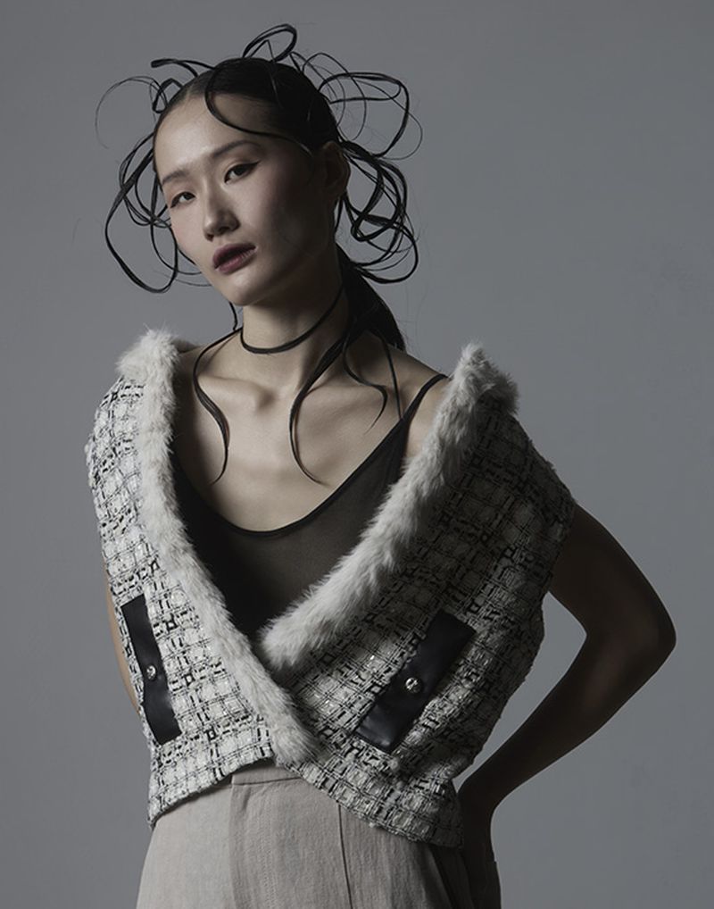 East Asian woman with sculptural tied hair wearing a tweed vest against a grey background