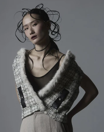 East Asian woman with sculptural tied hair wearing a tweed vest against a grey background