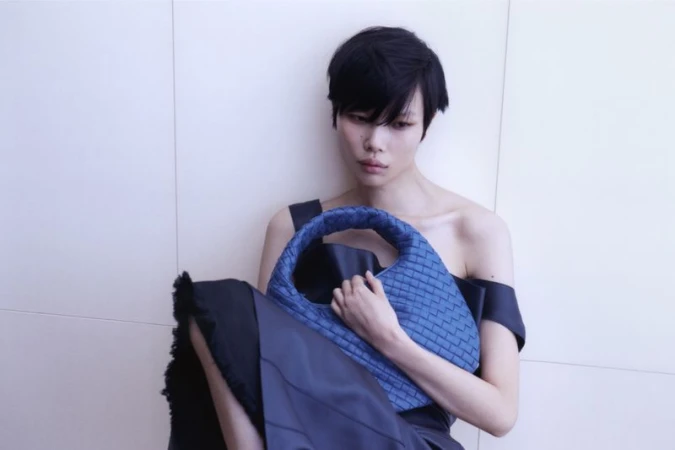Model with short dark hair holding a blue woven leather handbag against a plain light wall