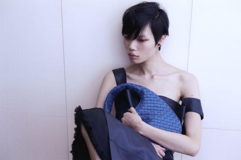 Model with short dark hair holding a blue woven leather handbag against a plain white wall