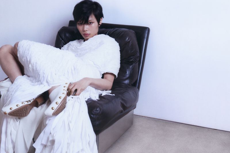 Model with short black hair seated in a dark leather armchair wearing white textured clothing against a plain background
