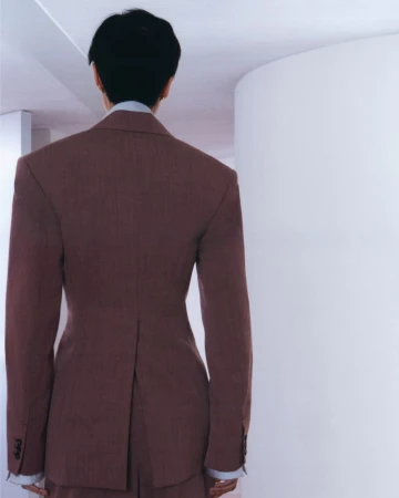 Model seen from behind wearing a structured brown blazer and light collared shirt against a white curved wall.