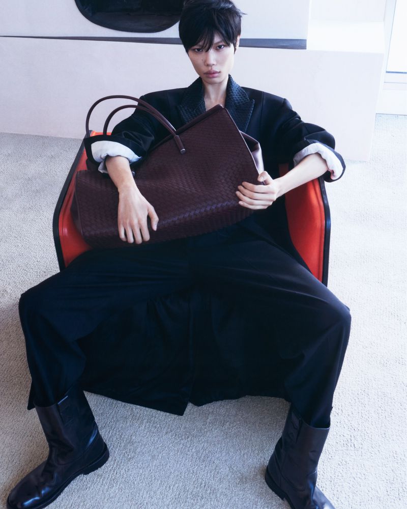 Woman in black blazer and trousers holding a woven leather purse while sitting in an orange and black chair in a well-lit room.
