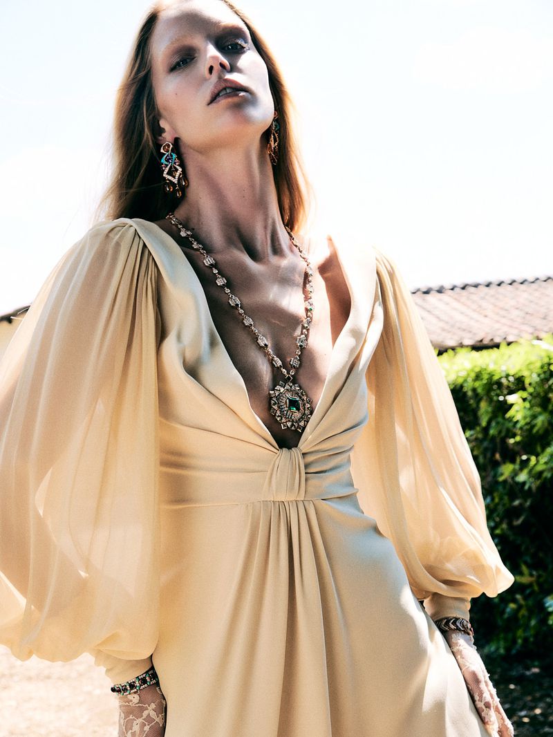 Fashion shot of a female model wearing a long-sleeved, pale yellow dress with a plunging neckline and jewelry outdoors.