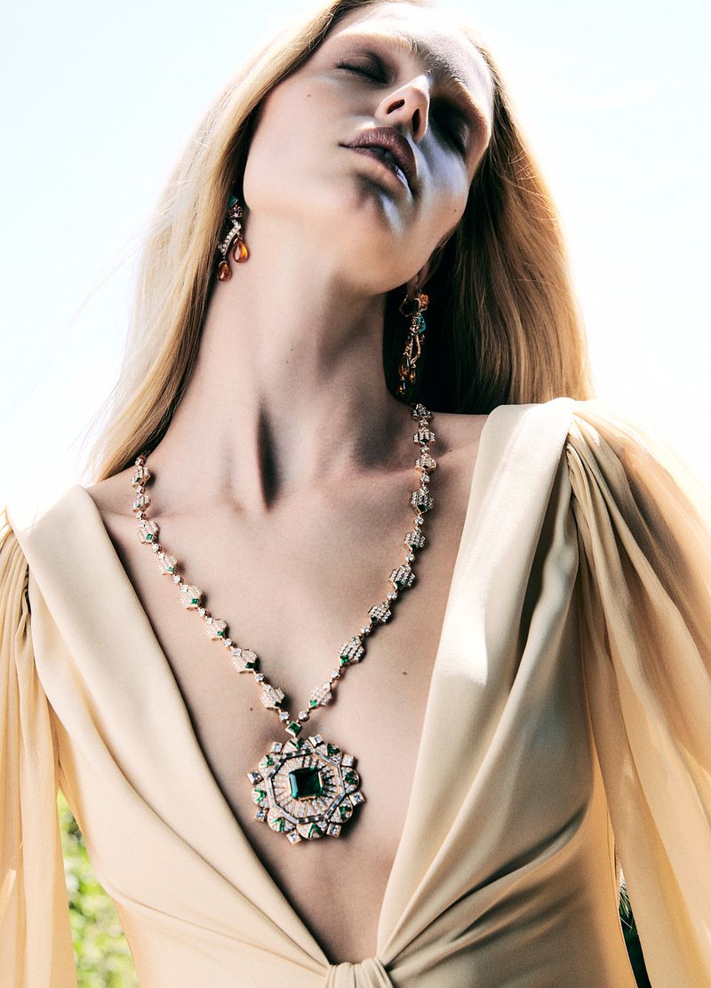 A blonde woman poses outdoors wearing a beige dress and jewelry, including a necklace, earrings, with natural light and a bright background.