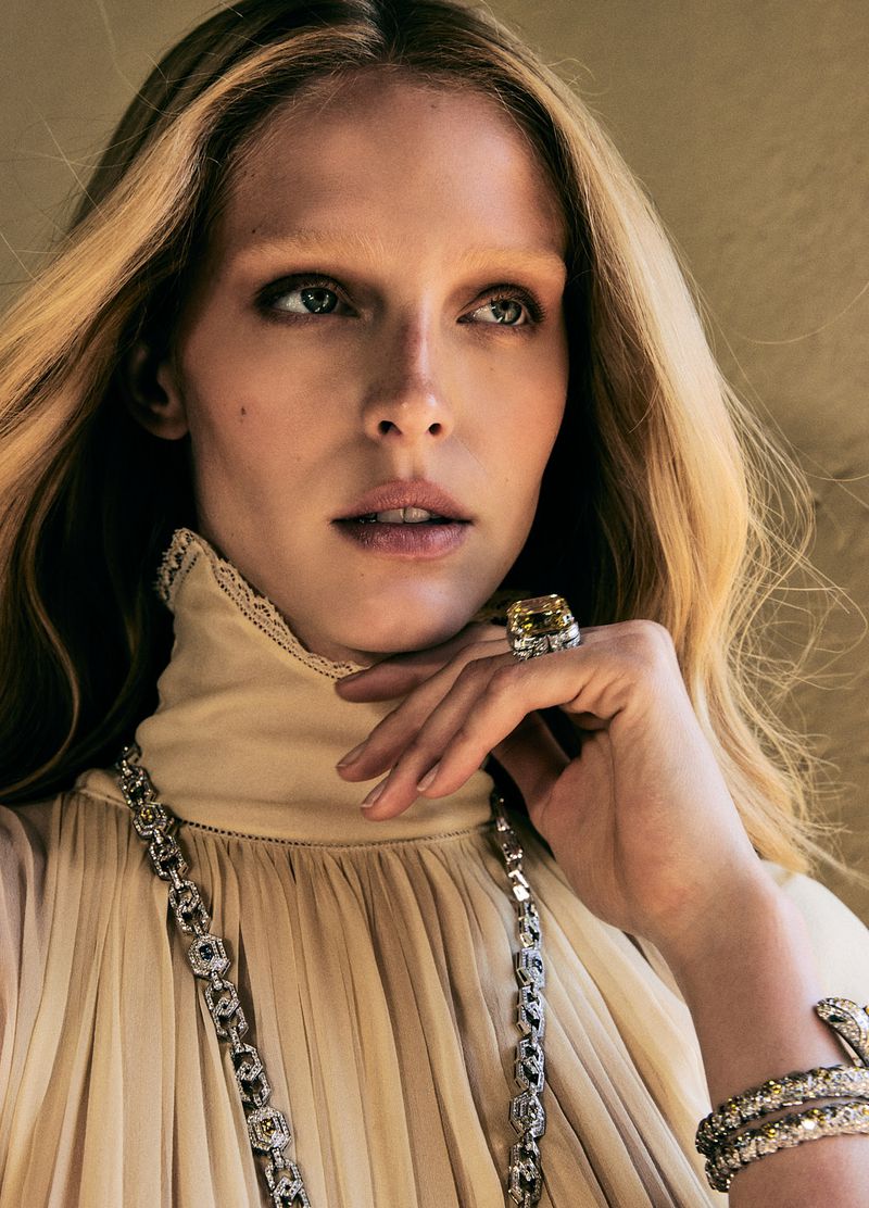 A woman with blonde hair, wearing a cream-colored, high-necked blouse and jewelry, poses in a close-up beauty shot. The background is blurred.