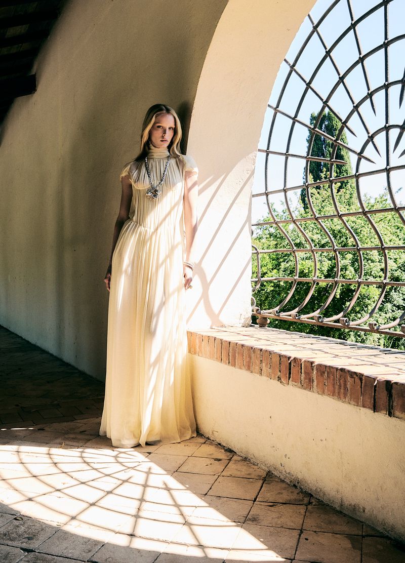 A woman in a long cream gown stands in a sunlit archway with decorative ironwork, highlighting fashion and architectural design.