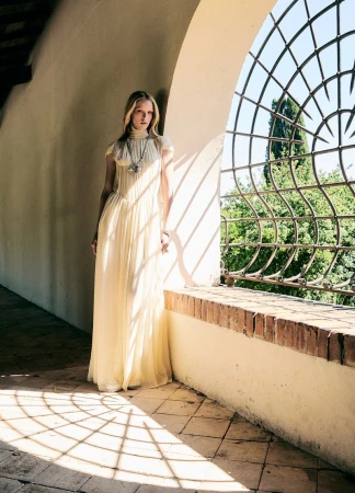A woman in a long cream gown stands in a sunlit archway with decorative ironwork, highlighting fashion and architectural design.