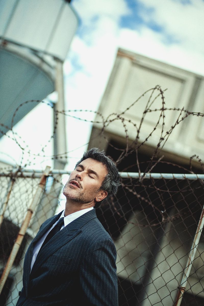 Man in dark pinstripe suit and tie posing outdoors against a chain-link fence with barbed wire and industrial structures