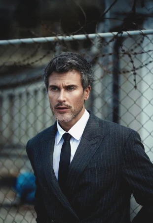 Close-up of a man with short grey-brown hair wearing a dark pinstripe suit, white shirt, and black tie against a chain-link fence.