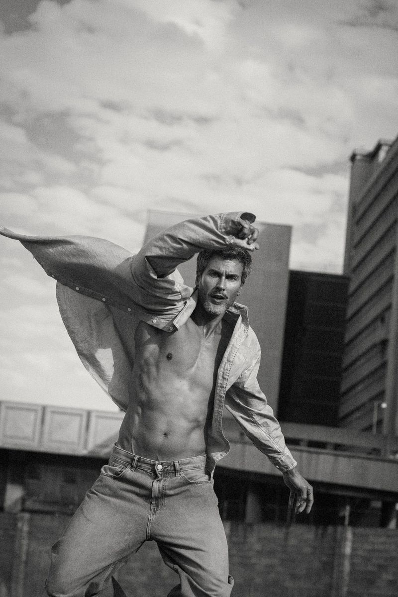 Monochrome fashion editorial portrait of a shirtless male model leaping outdoors with a billowing shirt against a cloudy sky and architectural background.