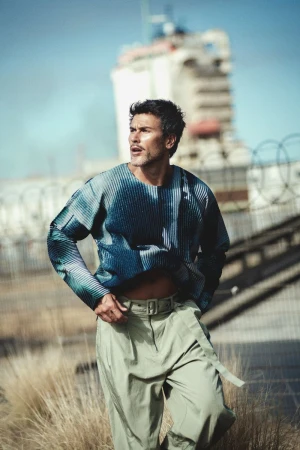 Man wearing a blue patterned long-sleeve top and light green belted trousers outdoors against a blurred industrial background.