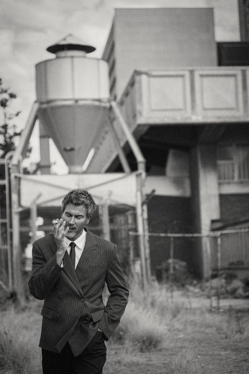 Monochrome portrait of a man in a double-breasted suit standing outdoors near industrial architecture.