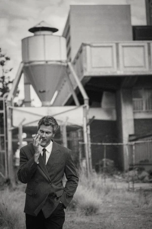 Monochrome portrait of a man in a double-breasted suit standing outdoors near industrial architecture.