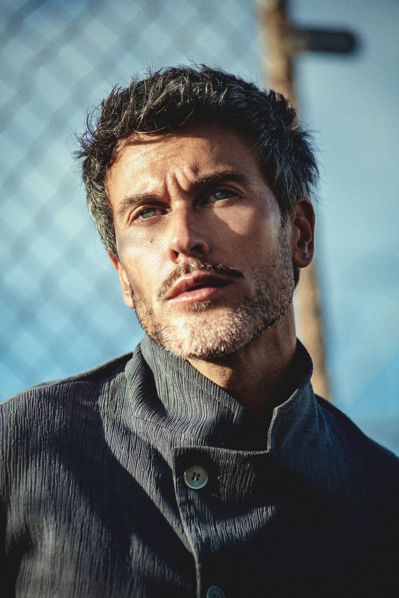 Portrait of a man with short dark hair wearing a high-collared dark jacket against a chain-link fence.