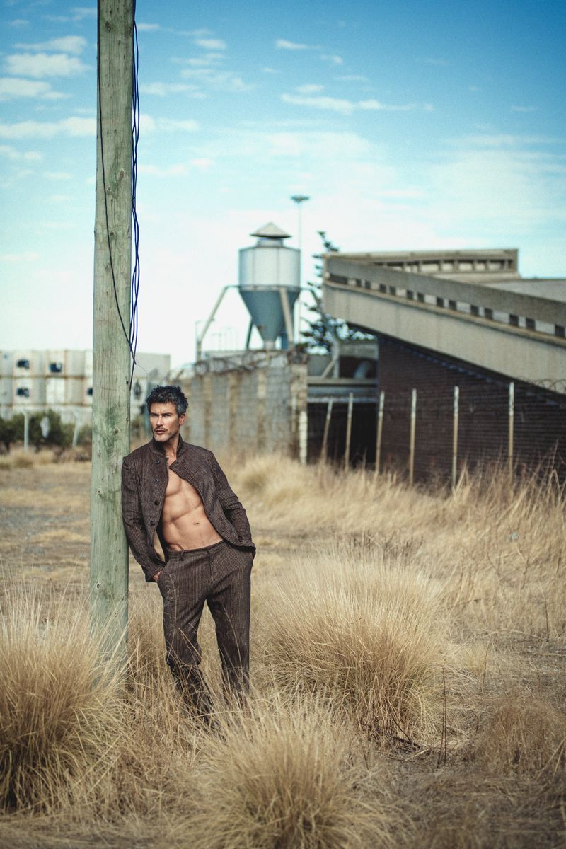 Shirtless male model leaning against a wooden utility pole in a dry grassy field with industrial structures behind.
