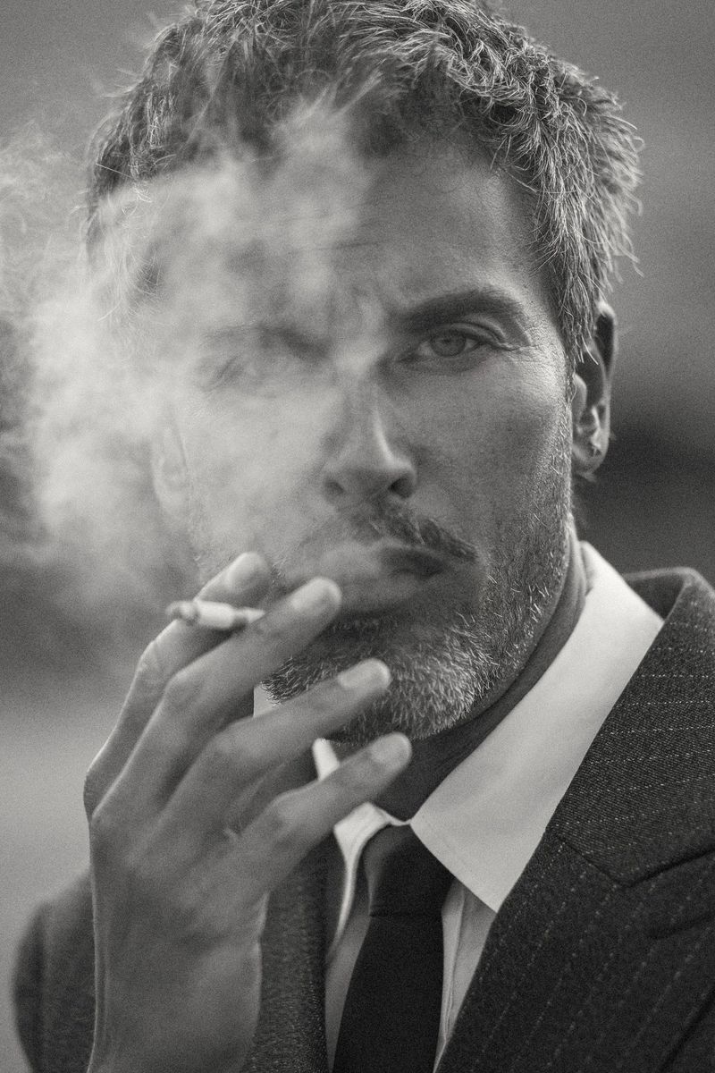 Monochrome portrait of a man with graying hair and mustache, wearing a suit and tie while smoking a cigarette with visible smoke.