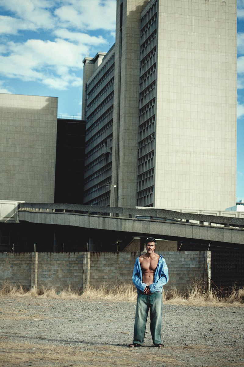 Shirtless man wearing loose denim jeans and an open denim shirt standing before a concrete wall and tall building