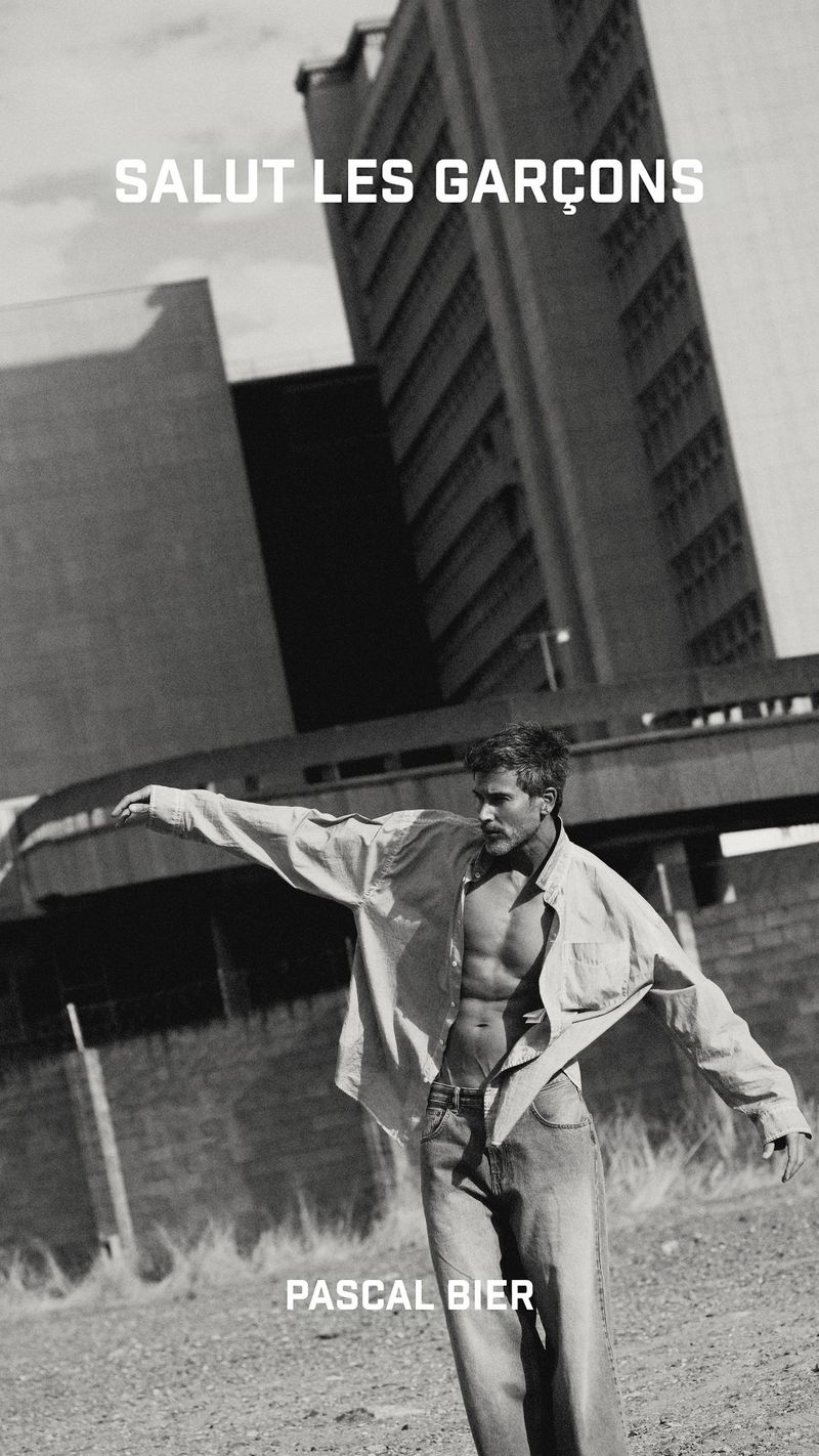 Black and white editorial image of a shirtless male model wearing loose trousers against a Brutalist building exterior.