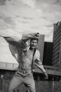 Monochrome fashion editorial portrait of a shirtless male model leaping outdoors with a billowing shirt against a cloudy sky and architectural background.