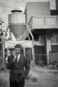 Monochrome portrait of a man in a double-breasted suit standing outdoors near industrial architecture.