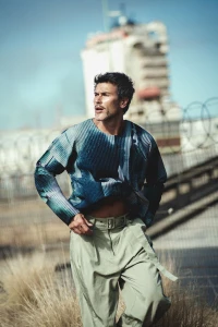 Man wearing a blue patterned long-sleeve top and light green belted trousers outdoors against a blurred industrial background.